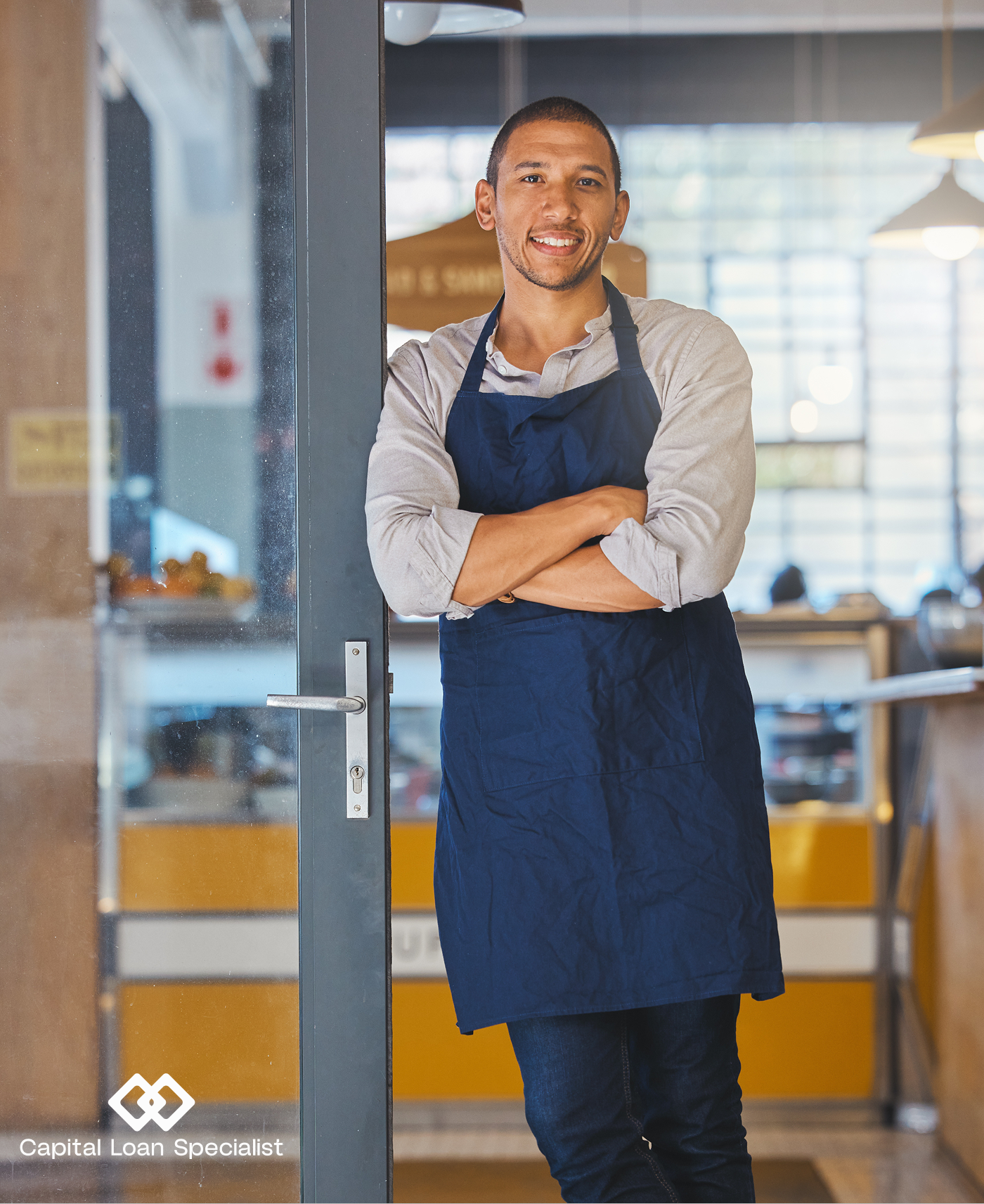 A smiling business owner wearing a blue apron, leaning against a glass door with a Capital Loan Specialist watermark.